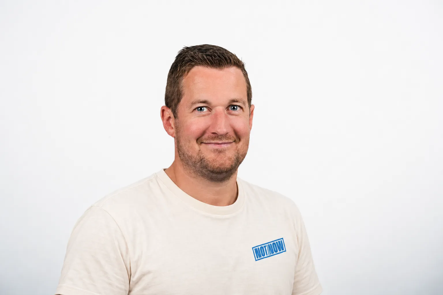 Glimlachende man met kort bruin haar en lichte baard, in gebroken wit T-shirt met blauw NOTNOW-logo.